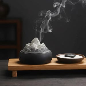 Peut inclure: Un brûleur d'encens en céramique noire avec un motif de montagne, dégageant des volutes de fumée blanche. Un petit plat rond avec du sable et une pierre sombre est posé à côté sur un plateau en bois. La scène évoque le calme et la tranquillité.