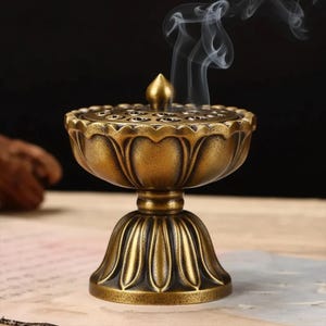Peut inclure: Un brûleur d'encens de couleur bronze avec un motif de fleur de lotus. Le brûleur a un bol rond avec de petits trous et un couvercle, reposant sur une base de piédestal. Des volutes de fumée s'élèvent du dessus.