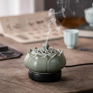 Peut inclure: Un brûleur d'encens en forme de fleur de lotus en céramique verte avec une base noire. Le brûleur émet de la fumée blanche. Une petite tasse à thé bleu clair est à l'arrière-plan. Le brûleur est sur une surface en bois.