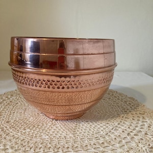 May include: A polished copper bowl with a smooth upper rim and a textured, hammered-style band around the middle section. The bowl features a rounded base and a reflective metallic finish that catches light, showcasing a warm, reddish-brown hue.