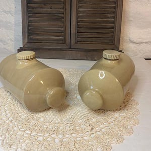 Può includere: Due borse dell'acqua calda cilindriche in gres beige con estremità arrotondate e tappi a vite. Ogni bottiglia presenta una finitura liscia e lucida e un'apertura centrale con un collo filettato, progettata per contenere liquidi.