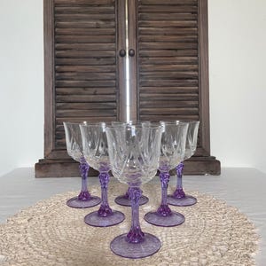 May include: Six clear crystal wine glasses with purple stems and bases are displayed on a cream lace doily. The glasses have a detailed, cut-glass design. A wooden shuttered door forms the background.