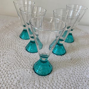 May include: Set of six clear glass cocktail glasses with teal-colored bases. The glasses have a conical bowl shape and a short stem. The glasses are arranged on a white, crocheted doily.