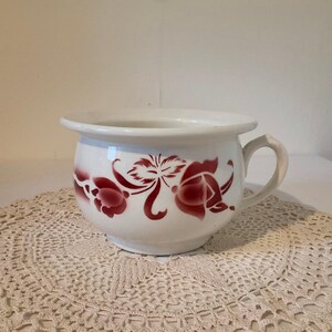 May include: A white ceramic chamber pot featuring a rounded body, a single side handle, and a wide rim. The exterior is decorated with a repeating floral pattern in a deep red color, showcasing stylized petals and leaves encircling the center.