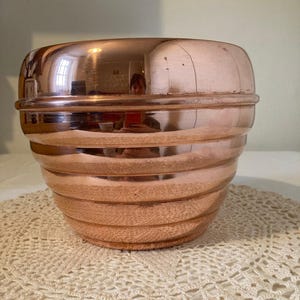 May include: A polished copper bowl with a rounded, tapered shape and horizontal ribbed detailing. The metallic surface is reflective, showing a distorted view of the surroundings. The bowl features a smooth, wide rim and a narrow base.