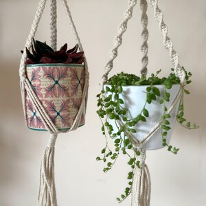 May include: Two macrame plant hangers with potted plants. One pot has a pink and blue floral design, the other is white. The hangers are made of cream-coloured rope and have a tassel at the bottom. The plants have green and burgundy leaves.