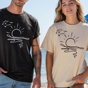 May include: A black t-shirt and a beige t-shirt, each featuring a minimalist line art graphic of a sun rising over waves with a flock of birds flying above. The black shirt has a white graphic, while the beige shirt has a black graphic.