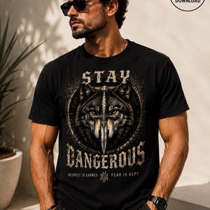 May include: A black short-sleeve t-shirt featuring a graphic of a wolf with a sword positioned between its eyes. The text reads STAY DANGEROUS with RESPECT IS EARNED FEAR IS KEPT printed below. The design is enclosed in a circular frame.