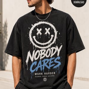 Peut inclure: T-shirt noir surdimensionné avec un motif vieilli. Le design présente un smiley blanc avec des X pour les yeux, les mots "NOBODY CARES" en blanc et "WORK HARDER. SILENCE. FOCUS. SUCCESS." en bleu. Un petit globe est en bas.