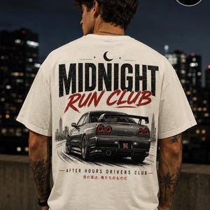 May include: An off-white t-shirt featuring a graphic on the back with a dark gray sports car, the text MIDNIGHT RUN CLUB, and the phrase AFTER HOURS DRIVERS CLUB with Japanese characters. A crescent moon icon sits above the bold black and red lettering.