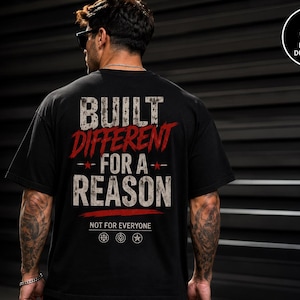 May include: A black short-sleeved t-shirt featuring the white and red distressed text BUILT DIFFERENT FOR A REASON, with the smaller text NOT FOR EVERYONE below it, along with three small circular icons featuring a barbell, a target, and a star.