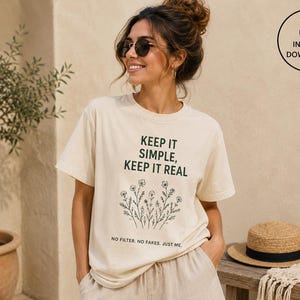 May include: Cream-colored t-shirt with the text "KEEP IT SIMPLE, KEEP IT REAL" in dark green, along with a floral illustration. The shirt also has the text "NO FILTER. NO FAKES. JUST ME." A straw hat with a black band sits on a wooden bench.