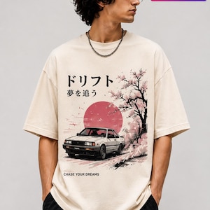 May include: Beige t-shirt with a Japanese design. It features a white car, a red sun, cherry blossoms, and Japanese text. The text "CHASE YOUR DREAMS" is printed on the shirt. A stylish tee.