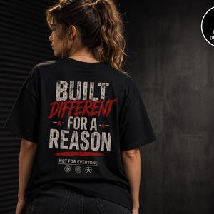 May include: A black short-sleeve t-shirt featuring the white and red distressed text BUILT DIFFERENT FOR A REASON, with the smaller text NOT FOR EVERYONE and three circular icons below, all printed on the back of the garment.