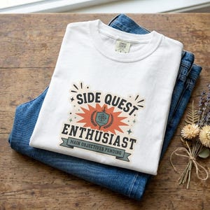 Puede incluir: Una camiseta blanca de manga corta con un estampado gráfico que dice SIDE QUEST ENTHUSIAST y MAIN OBJECTIVES PENDING, centrado alrededor de un icono de escudo y espada con un fondo de estallido estelar, doblada sobre unos vaqueros azules.