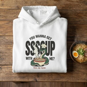 May include: White hoodie with a graphic of a snake chef with the text "YOU WANNA GET SSSOUP WITH ME?" and "hisss. sip. repeat." A bowl of ramen and chopsticks are next to the hoodie on a wooden table.