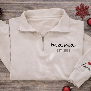 Könnte beinhalten: Ein cremefarbenes Sweatshirt mit einem Kragen mit Viertelreißverschluss. Auf der Vorderseite stehen die Worte "mama" und "EST. 2026". Der Ärmel hat den Namen "Zara" und ein rotes Herz. Das Sweatshirt liegt auf Holz mit Weihnachtsdekoration.