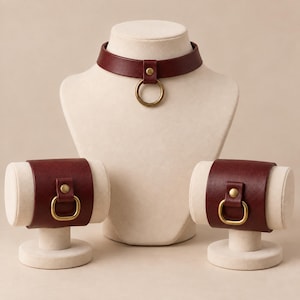 Könnte beinhalten: Ein dunkelrotes Lederhalsband und zwei passende breite Lederarmbänder. Jedes Teil hat eine glatte, matte Oberfläche, eine mittige Niete in Messingoptik und einen runden Metallring in Messingoptik, der an der Vorderseite befestigt ist.