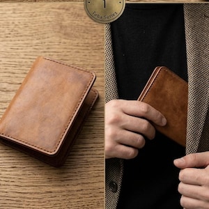 Può includere: Un portafoglio in pelle marrone con un bordo cucito è appoggiato su una superficie di legno. Il portafoglio è rettangolare e sembra nuovo. Una persona viene mostrata mentre mette un portafoglio simile nella tasca della giacca. Il portafoglio è di colore marrone chiaro.