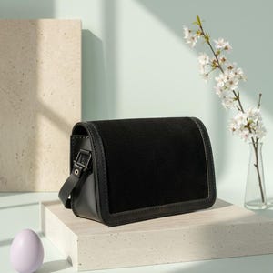 Peut inclure: Un sac noir en bandoulière de forme rectangulaire avec une fermeture à rabat. Le sac est doté d'un panneau en daim et d'une bandoulière en cuir. Un petit œuf violet clair et un vase de fleurs blanches sont également dans l'image.