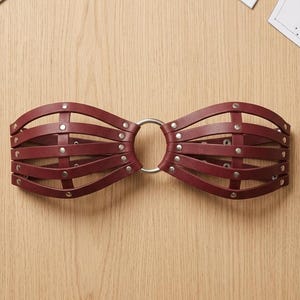 Puede incluir: Un cinturón de cuero rojo oscuro estilo jaula con múltiples correas horizontales conectadas por remaches de metal plateado y un anillo central de metal plateado. El diseño consta de dos secciones simétricas unidas en el centro.