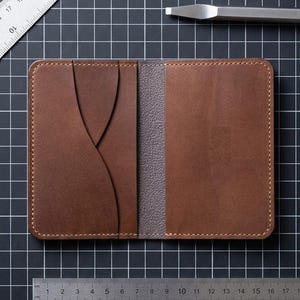 Può includere: Un portafoglio in pelle marrone con fessure per carte, posto su una superficie a griglia nera con strumenti per la lavorazione della pelle e un righello. Il portafoglio è aperto, rivelando un interno grigio. Un righello di metallo mostra le misure in cm.