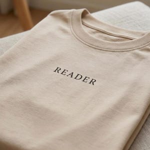 Könnte beinhalten: Beiges T-Shirt mit dem Wort "READER" in Schwarz auf der Brust gedruckt. Das Shirt ist sauber gefaltet und zeigt seinen Rundhalsausschnitt und den weichen Stoff. Die neutrale Farbe und das schlichte Design deuten auf einen lässigen Stil hin.