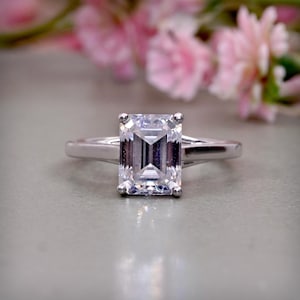 Könnte beinhalten: Ein silberfarbener Ring mit einem großen, klaren, im Smaragdschliff geschliffenen Mittelstein, der von vier Krappen gehalten wird. Die Ringschiene ist glatt und poliert und weist ein geteiltes Design auf, das in die Fassung übergeht.