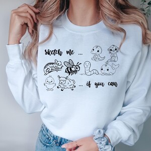 Könnte beinhalten: Weißes Sweatshirt mit einer schwarzen Strichzeichnung verschiedener Tiere, darunter ein Dinosaurier, eine Biene und ein Wal. Der Text "sketch me... if you can" ist auf der Vorderseite aufgedruckt.