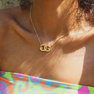 Puede incluir: Un collar dorado con un colgante de doble letra. El colgante tiene un diseño estilizado y la cadena es fina. El collar se lleva sobre la piel, con un top colorido visible en el fondo.