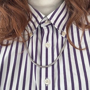 May include: A silver chain necklace on a white and purple striped shirt. The shirt is partially unbuttoned, revealing a grey turtleneck. Red hair is visible at the top of the image.