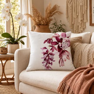 Può includere: Un cuscino quadrato bianco con un motivo floreale ad acquerello nei toni del bordeaux e del rosa. Il cuscino è su un divano beige, con altri cuscini decorativi e una coperta color terracotta. Un'orchidea in vaso è visibile sullo sfondo.