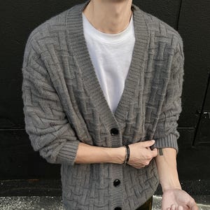 May include: A gray, textured cardigan sweater with a V-neck and black buttons. The sweater is worn over a white t-shirt. The cardigan has a woven pattern and long sleeves. A black bracelet is visible on the wrist.