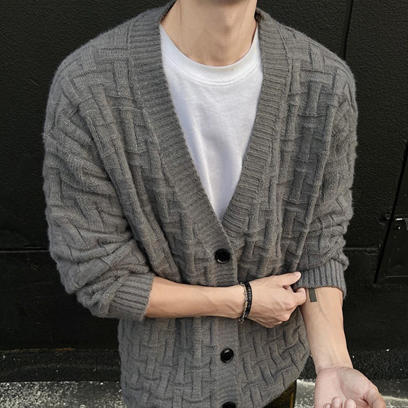 May include: A gray, textured cardigan sweater with a V-neck and black buttons. The sweater is worn over a white t-shirt. The cardigan has a woven pattern and long sleeves. A black bracelet is visible on the wrist.