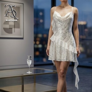May include: A white sequined mini dress with spaghetti straps and a cowl neckline. The dress features a ruffled accent on the side and a flowing fabric detail. The image also shows a glass of champagne on a glass table.