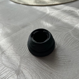 Könnte beinhalten: Ein schwarzes, zylindrisches Objekt mit strukturierter Außenseite und geriffeltem Boden. Das Objekt hat eine hohle Mitte und scheint aus einem festen Material zu bestehen. Der Artikel liegt auf einer weißen, gemusterten Oberfläche.