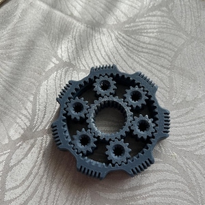 Può includere: Un meccanismo a ingranaggi grigio stampato in 3D con più ingranaggi interconnessi. L'ingranaggio centrale è circondato da ingranaggi più piccoli, il tutto all'interno di un ingranaggio esterno più grande. Il design è intricato e dettagliato, con un tema meccanico.