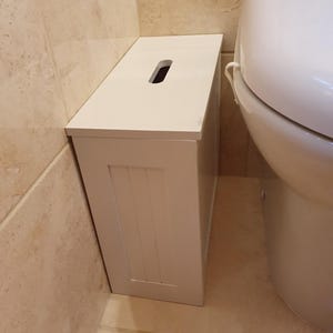 May include: A white bathroom storage cabinet, rectangular in shape, with a recessed handle on the top. The cabinet features a vertical panel design on the front and is placed beside a white toilet. The cabinet is made of wood.