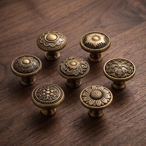 May include: A collection of seven round cabinet knobs featuring antique brass finishes and varied embossed patterns. Each knob displays distinct circular designs, including floral motifs, geometric shapes, and intricate scrollwork, all set on a central pedestal base.