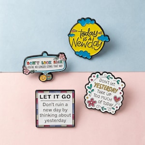 Puede incluir: Cuatro pines de esmalte con citas inspiradoras. Uno dice "Today is a Newday" en amarillo y azul. Otro dice "Don't Look Back" con detalles florales. Un tercero dice "Let It Go". El último dice "Don't let Yesterday Take Up Too Much of Today".
