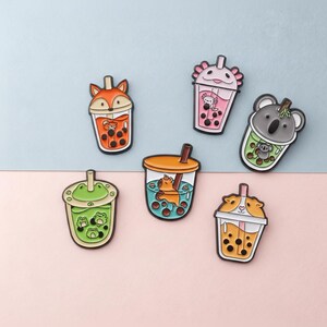May include: A collection of six colorful bubble tea-themed pins. Each pin features a different animal design, including a fox, axolotl, koala, frog, bear, and hamster. The pins are set against a light blue and pink background.