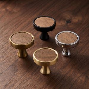 Puede incluir: Cuatro pomos circulares para armarios con una incrustación central de madera marrón natural. Los pomos tienen cuatro acabados metálicos distintos: negro mate, latón cepillado, cromo pulido y oro satinado, cada uno con una base ensanchada.