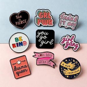 May include: A collection of enamel pins with various designs and text. The pins include phrases like "She is Fierce," "GRL PWR," "Shake it Off," "Be Kind," "You Go Girl," "Girl Gang," "Drama Queen," "Work Hard Dream Big," and "A Hug From Mum."