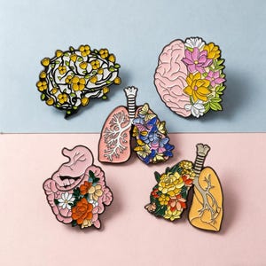 May include: A collection of enamel pins featuring anatomical designs with floral accents. The pins include a brain, lungs, and intestines, each adorned with colorful flowers. The pins are set against a light blue and pink background.