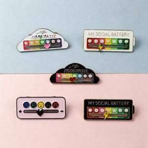 May include: Enamel pins with mood tracker and social battery designs. The pins feature a range of emotions, from sad to happy, and include text like "Moodtracker" and "My Social Battery". The pins are in various shapes and colors.