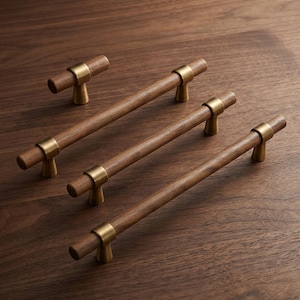 May include: A collection of four cabinet pulls featuring cylindrical dark brown wood bars accented with brushed brass metal mounting posts. The set includes one small T-shaped knob and three longer horizontal handles, each with two brass supports.