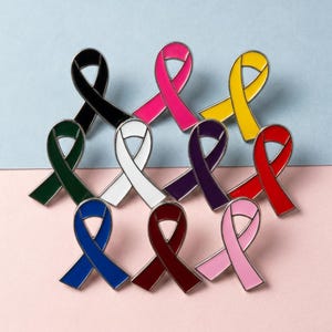 May include: A collection of enamel ribbon pins in various colors, including black, pink, yellow, red, and blue. Each pin has a silver-colored border and is arranged on a two-tone pink and blue background. These pins are a symbol of awareness.