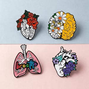 May include: Four enamel pins with anatomical designs. One pin features a heart with roses and the words "Heart Blood". Another shows a brain with daisies and bees. A third pin depicts lungs with the word "breathe". The last pin is a heart with flowers.