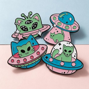 May include: Four enamel pins featuring cat-alien hybrids in UFOs. The pins have a black outline with pink, blue, green, and white colors. The cat-aliens have three eyes and are surrounded by stars and planets.