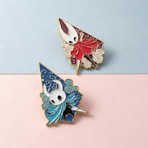 May include: Two enamel pins featuring video game characters. One pin has a red cape and white mask, the other has a blue cape and white mask. Both pins have gold-coloured metal accents and floral details.